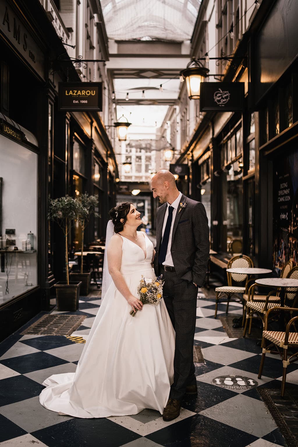A newlywed couple share an affectionate gaze in a charming vintage-style arcade. the bride in a white gown and the groom in a gray suit clasp hands, surrounded by quaint shopfronts and bistro chairs.