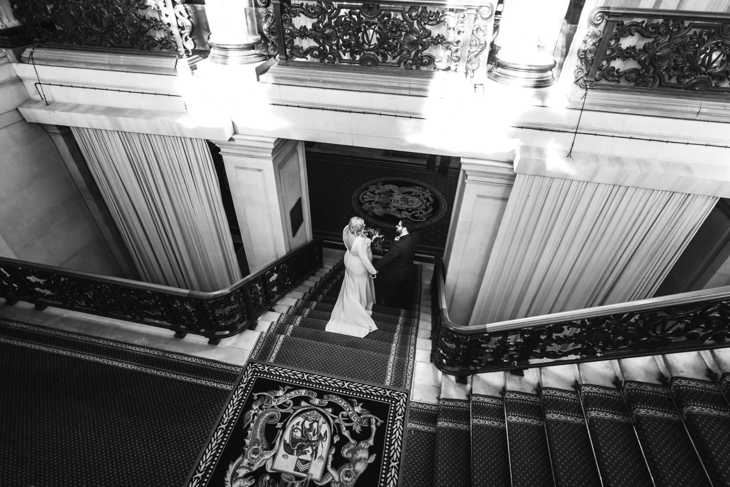 A bride and groom hold hands in a majestic stairwell, surrounded by elegant marble columns and intricate iron railings, with sunlight streaming through large windows onto the ornate floor.