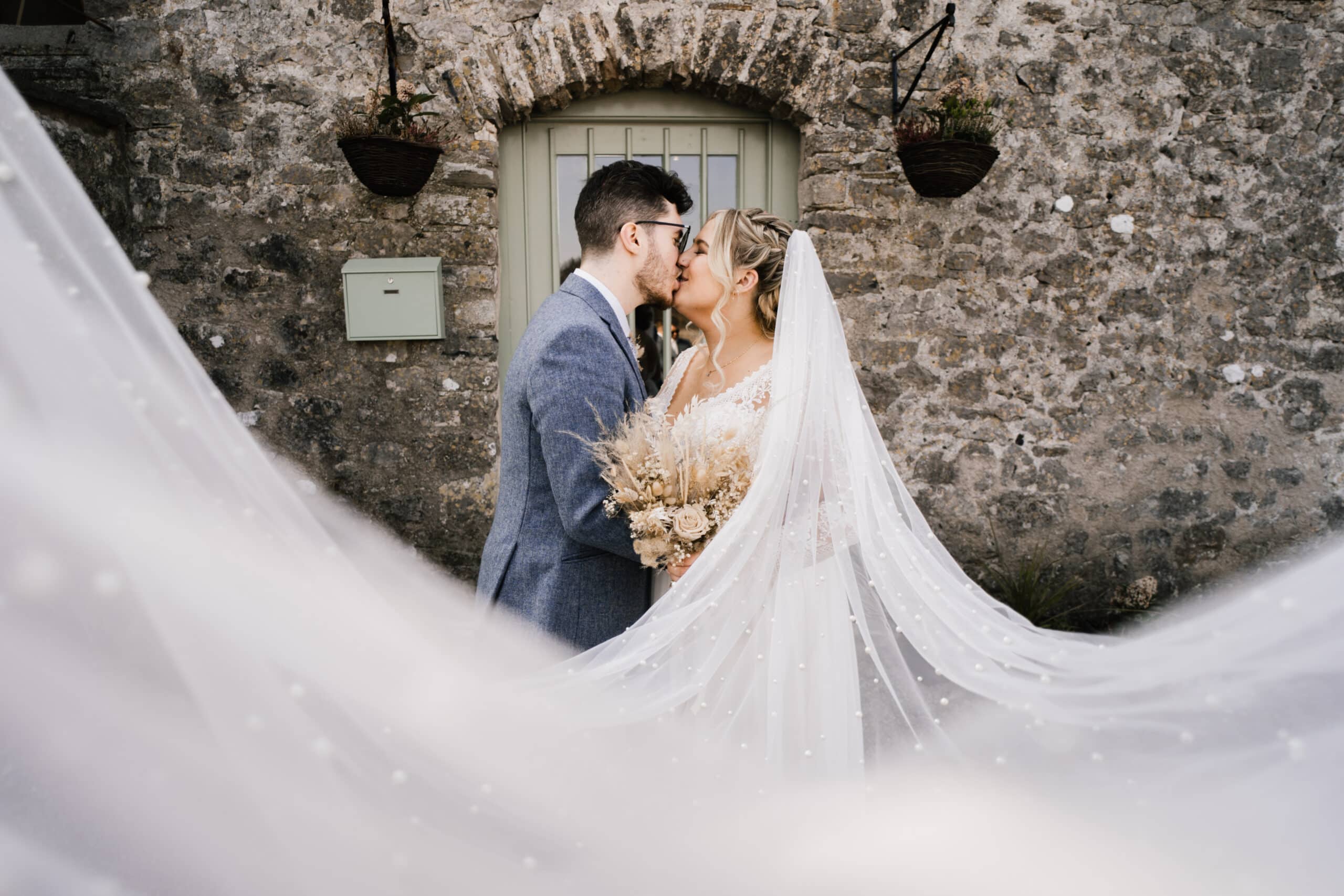 A bride and groom kiss outside a stone building, framed by the bride's flowing veil. she holds a bouquet, and hanging plants decorate the wall behind them.
