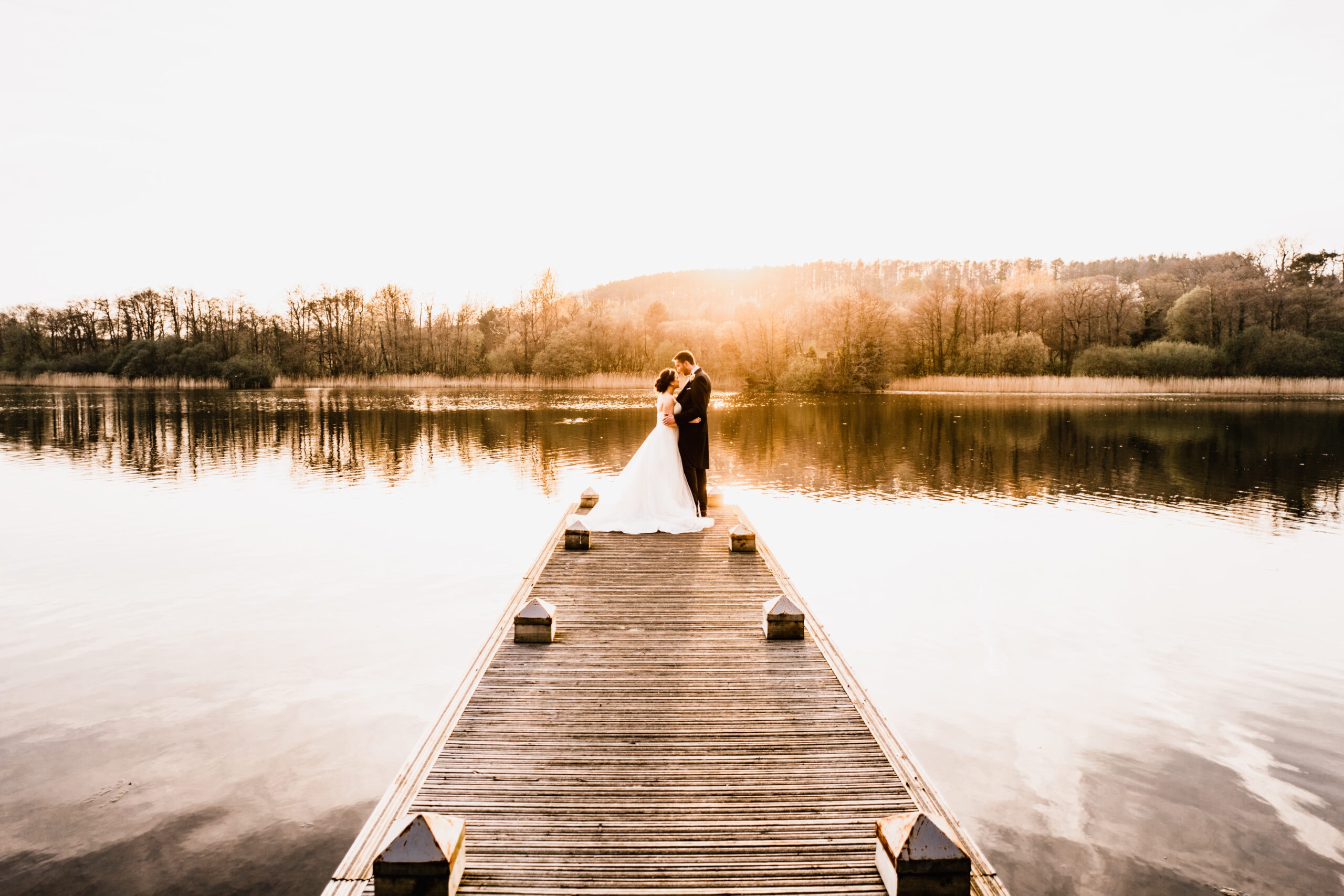 A bride and groom embrace at the end of a wooden dock, overlooking a serene lake at sunset, with warm sunlight casting a golden glow over the scene.
