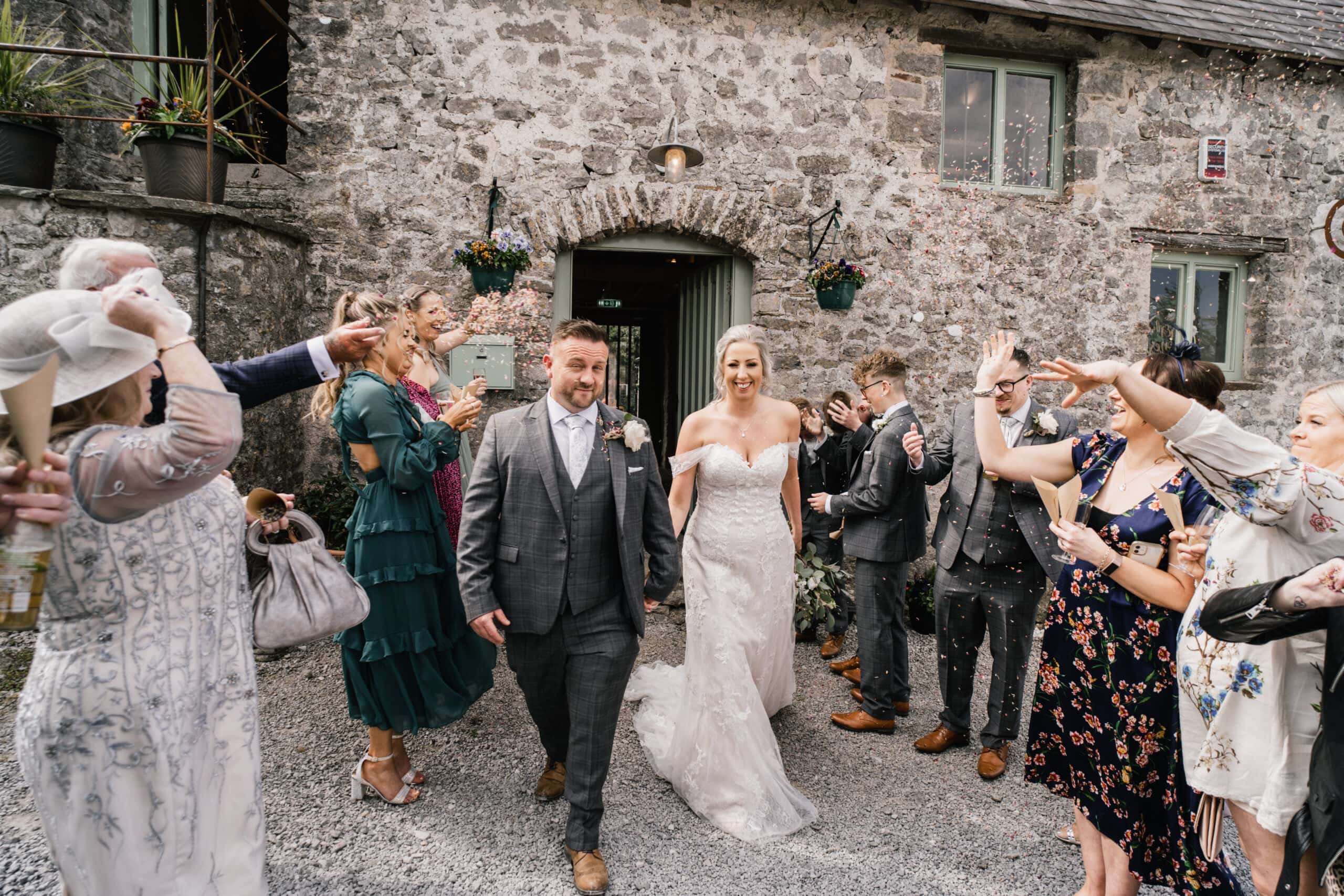 A joyful bride and groom exit a rustic stone building, smiling as guests line up on each side, tossing flower petals and raising glasses to celebrate.