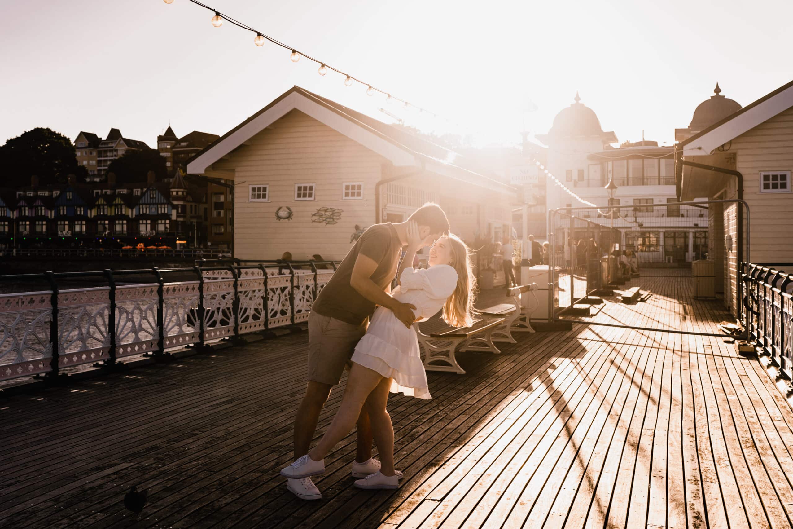 A couple kisses tenderly on a wooden pier at sunset, with soft golden light illuminating the scene and quaint buildings in the background.