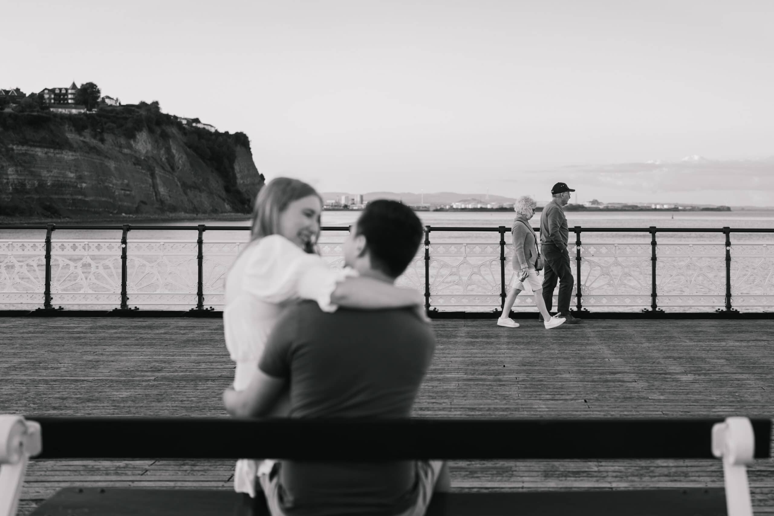 A black and white photo showing a couple embracing and smiling at each other on a pier, with an elderly couple walking in the background and a seascape with cliffs visible under a clear sky.