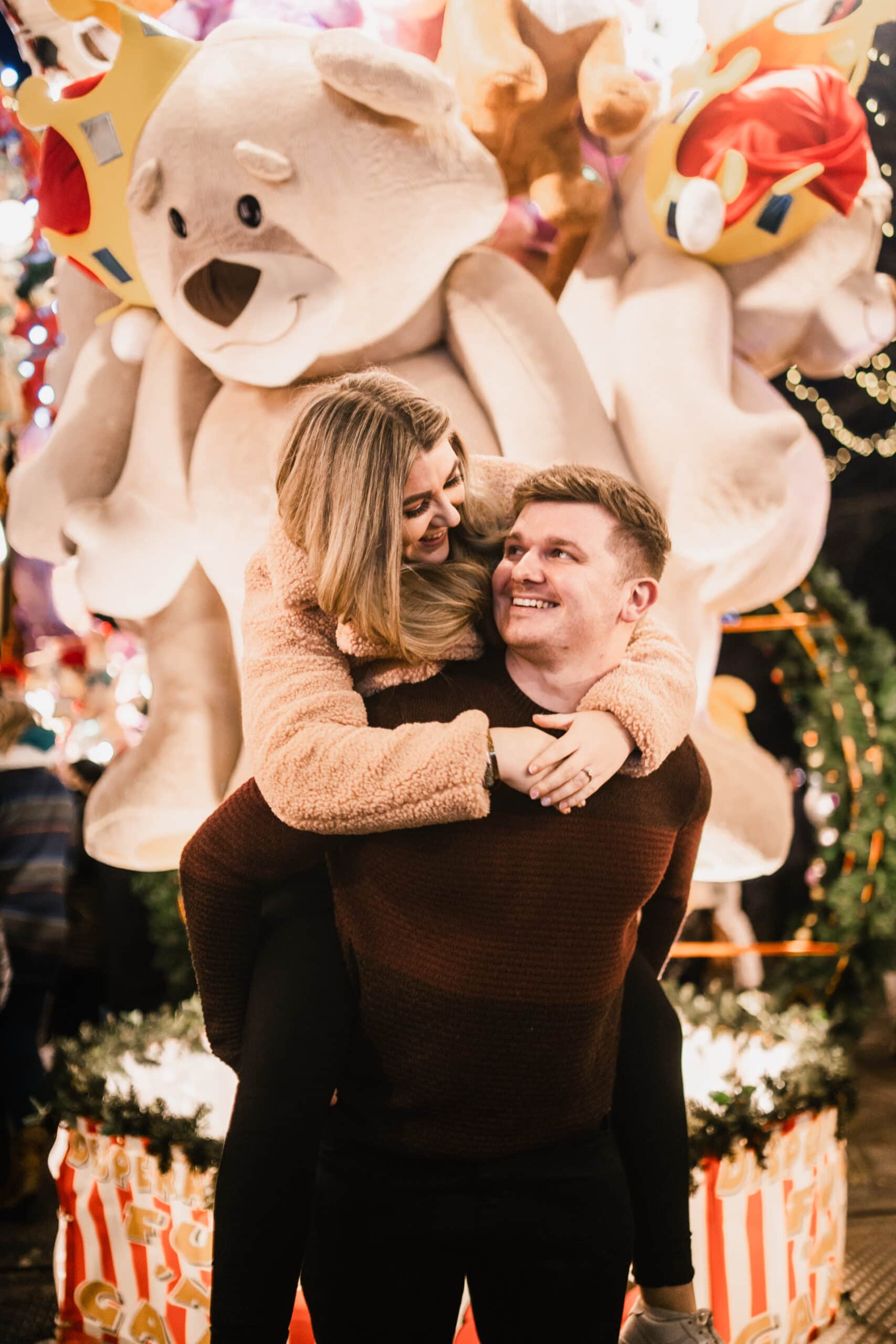 A joyful couple embraces in front of a festive backdrop with large plush toys and christmas decorations, sharing a tender moment under glowing lights.
