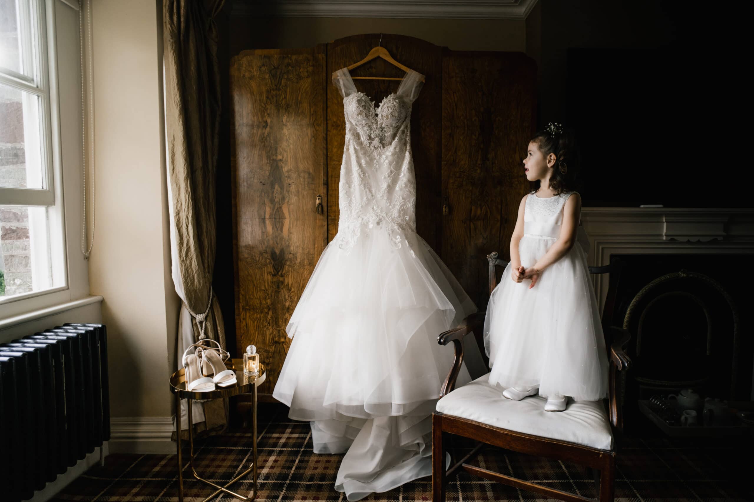 A young girl in a white dress gazes at an elegant wedding gown hanging on a wooden wardrobe in a vintage-styled room, natural light streaming through the window, capturing the essence of wedding photography style
