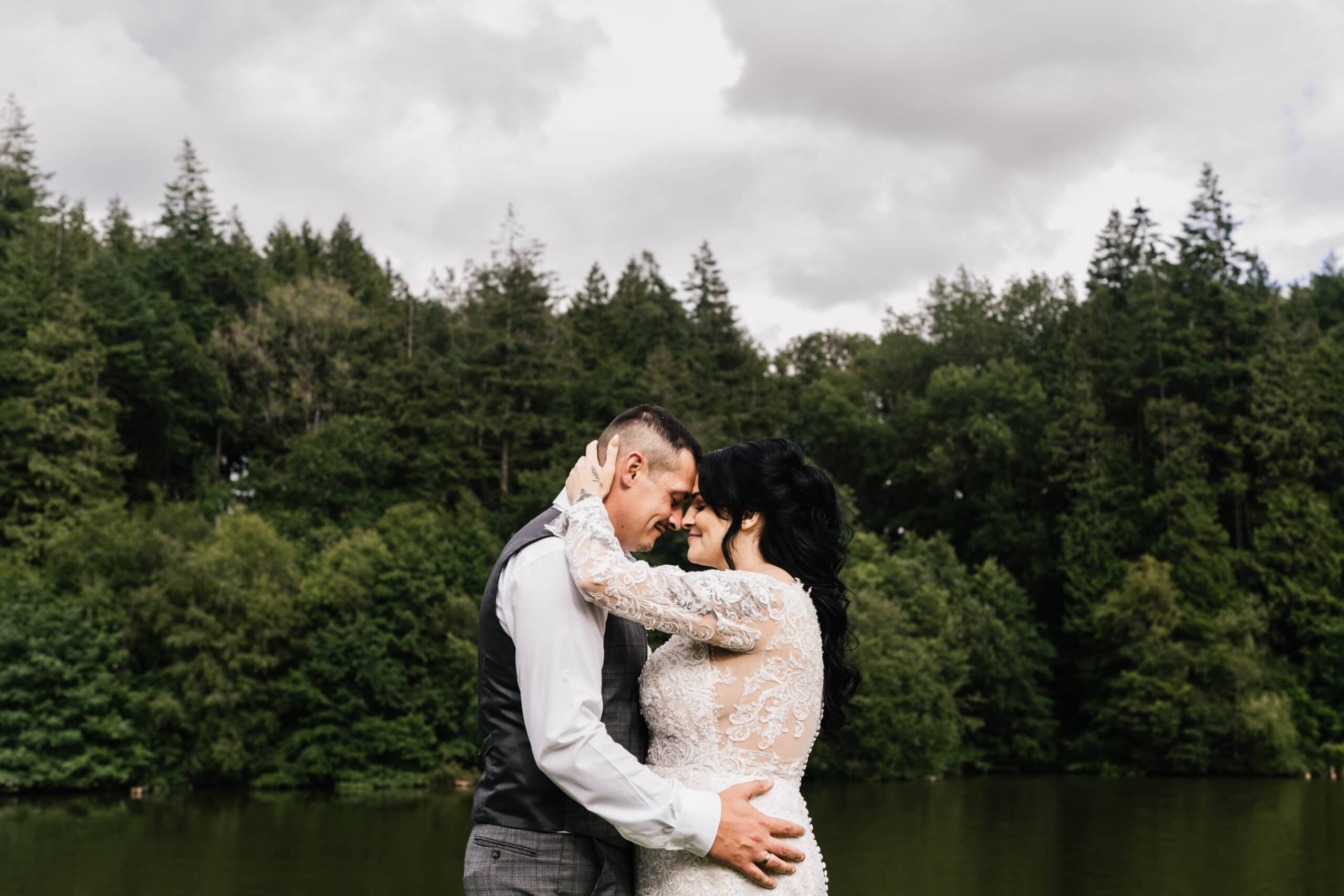 A couple embraces, foreheads touching, beside a serene lake at a Canada Lodge with a dense forest in the background under a cloudy sky. The woman wears a lace dress and the man is in a