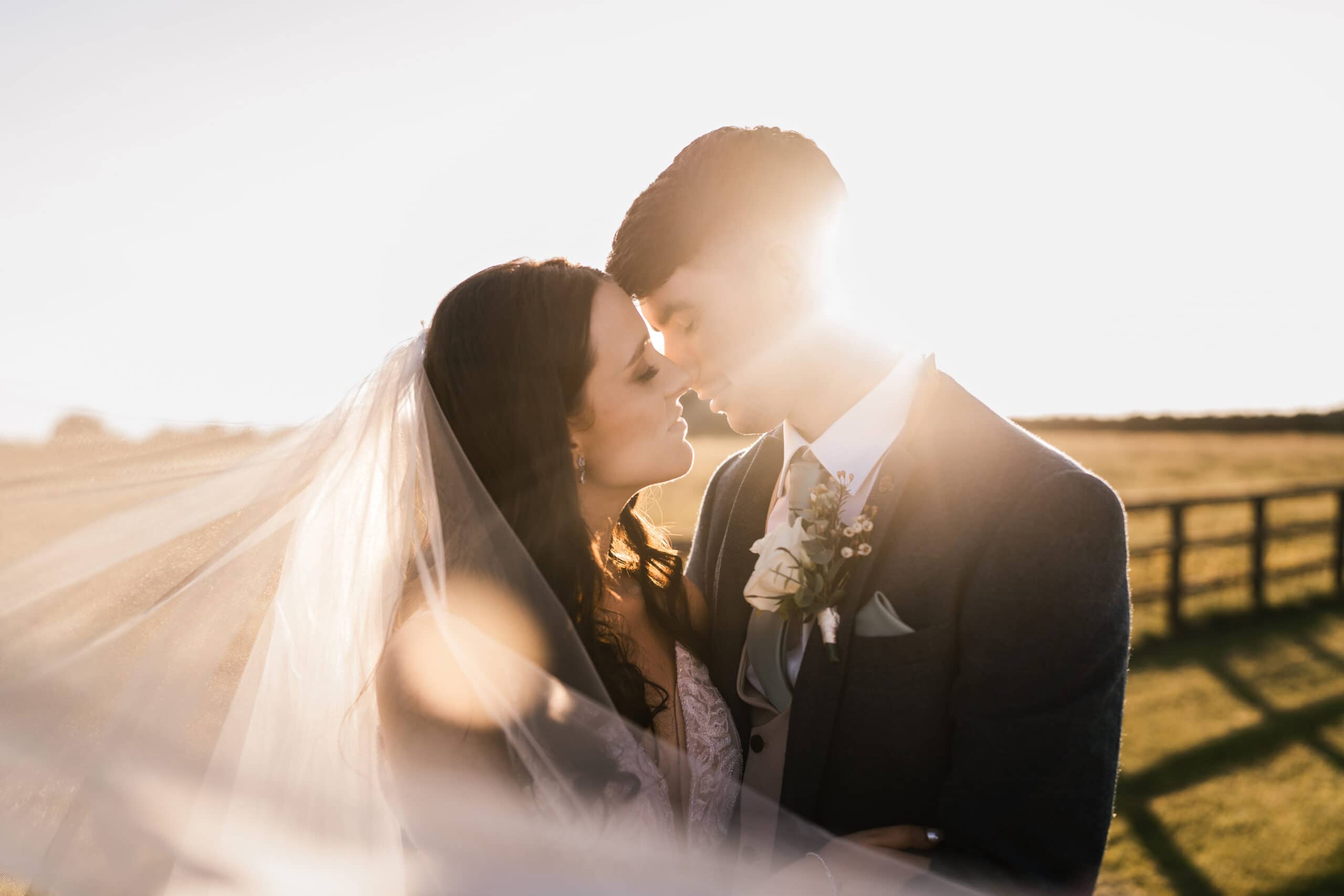 A newlywed couple sharing a romantic moment in a sunlit field, with the bride's veil and the groom's suit highlighted by the sunset.