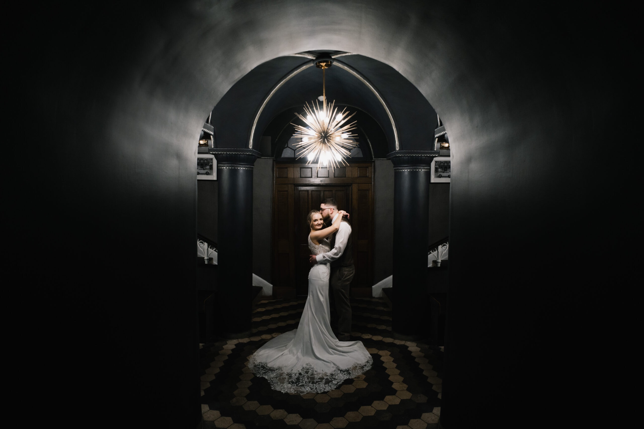 A newlywed couple embraces under a striking light fixture in a dark, arched hallway with a patterned floor, creating a dramatic and romantic scene.