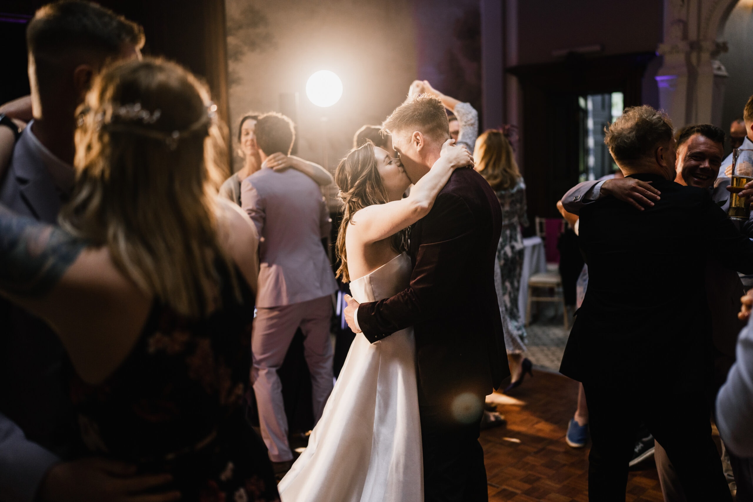 A bride and groom share a romantic kiss on the dance floor surrounded by guests dancing and celebrating at a lively wedding reception.
