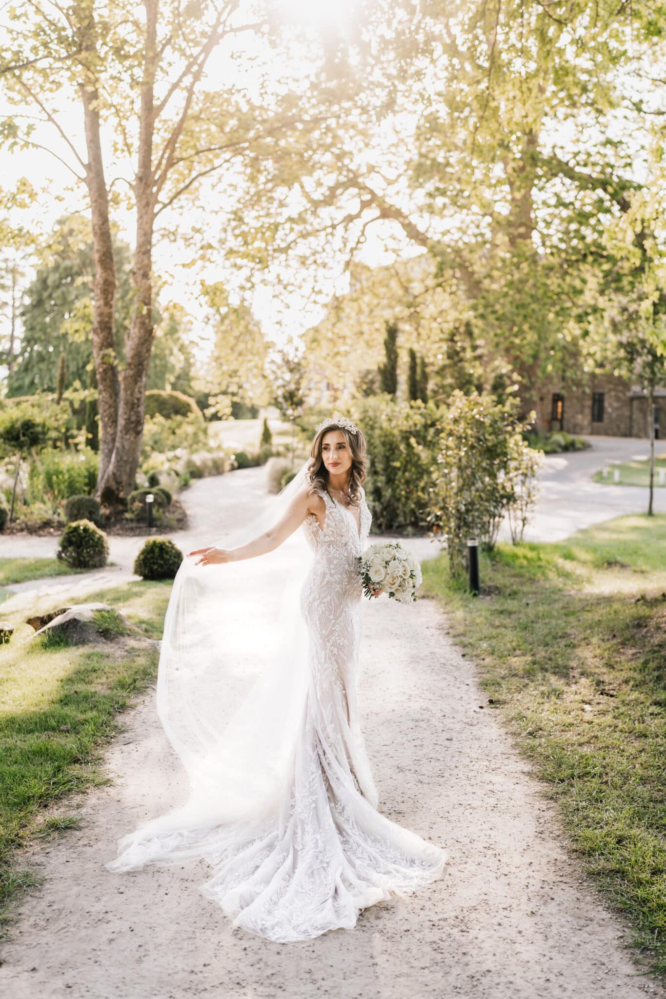 A bride in a detailed lace gown holds her veil flowing in the breeze, standing on a garden path with sunlight filtering through trees. she holds a bouquet, looking over her shoulder.