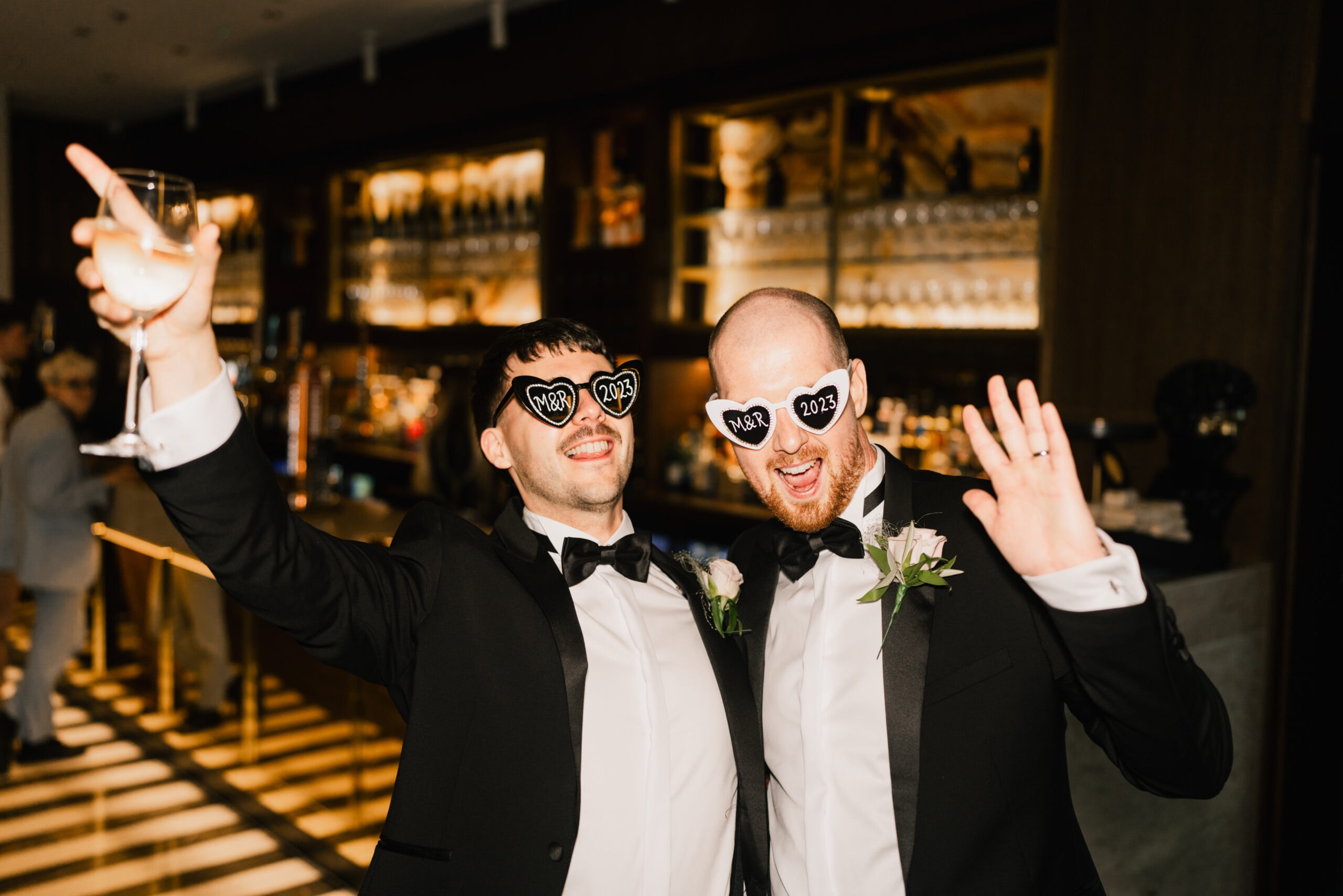 Two men in tuxedos at a celebration, wearing sunglasses shaped like "2024". they are smiling and taking a selfie, standing in a warmly lit venue with a bar in the background.