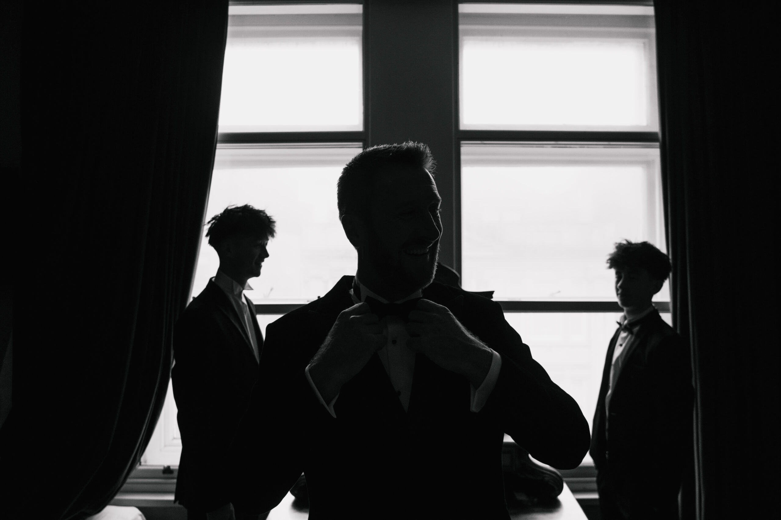 A man in a suit adjusts his bow tie, silhouetted against a bright window, with two other men in the background. The mood is formal and elegant, typical of a South Wales wedding