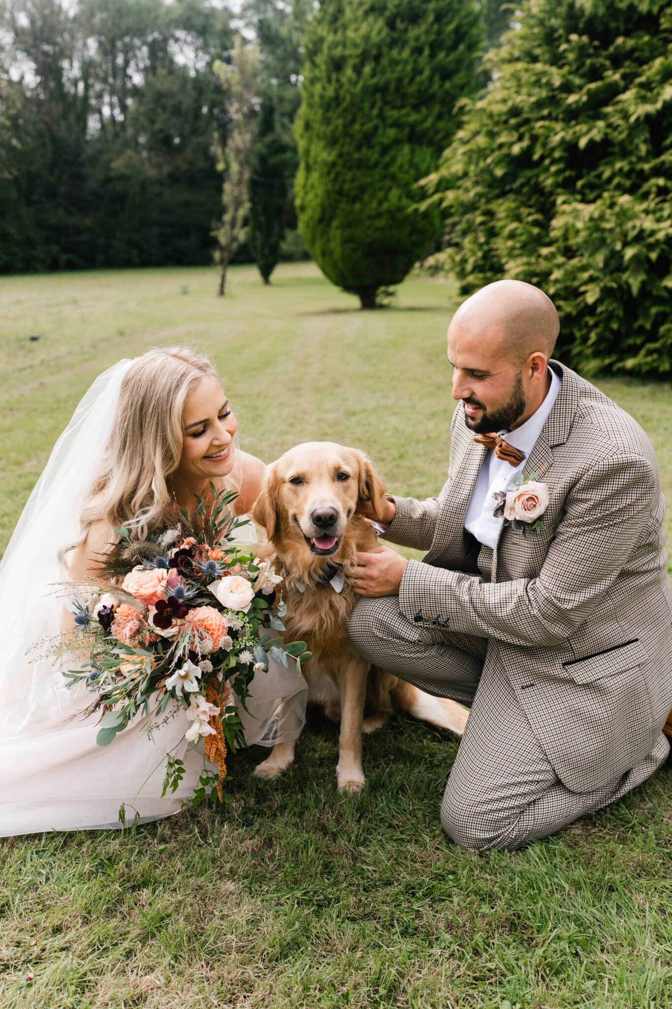 A bride and groom happily petting their golden retriever in a lush garden in South Wales, the bride holding a large floral bouquet, both dressed in wedding attire.