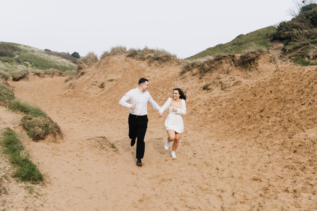 A joyful couple runs hand in hand along a sandy path surrounded by grassy dunes, both dressed in white, with bright, cheerful expressions.