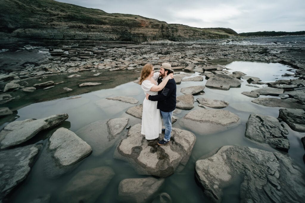 A couple stands embracing on large rocks surrounded by shallow water and rocky terrain, with hills in the background under a cloudy sky.