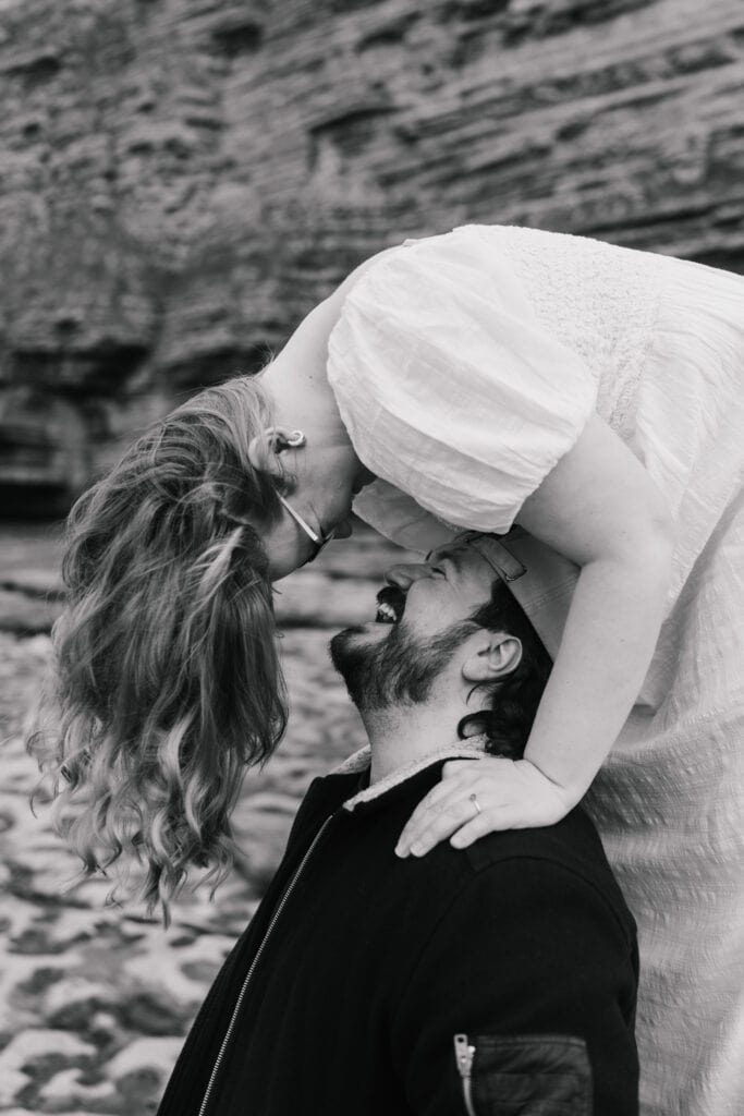 A woman in a white dress leans upside down towards a seated man, their faces close and smiling, against a rocky outdoor background. The scene is joyful and intimate, captured in black and white.