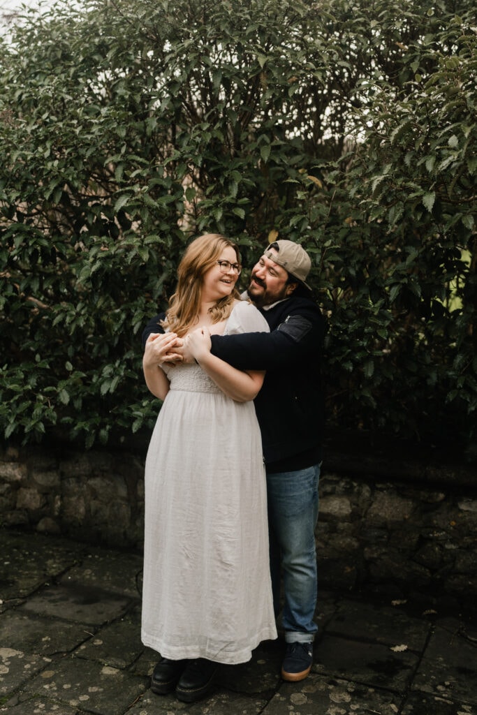 A man in a cap embraces a smiling woman in a white dress from behind. They stand on a stone path in front of dense green bushes, looking at each other happily.