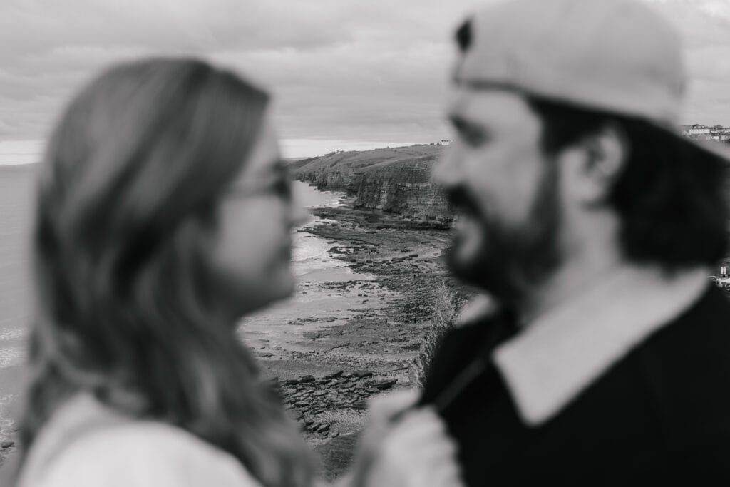 A black and white photo of two people facing each other in the foreground, out of focus, with a sharp, detailed coastline and sea cliffs in the background.