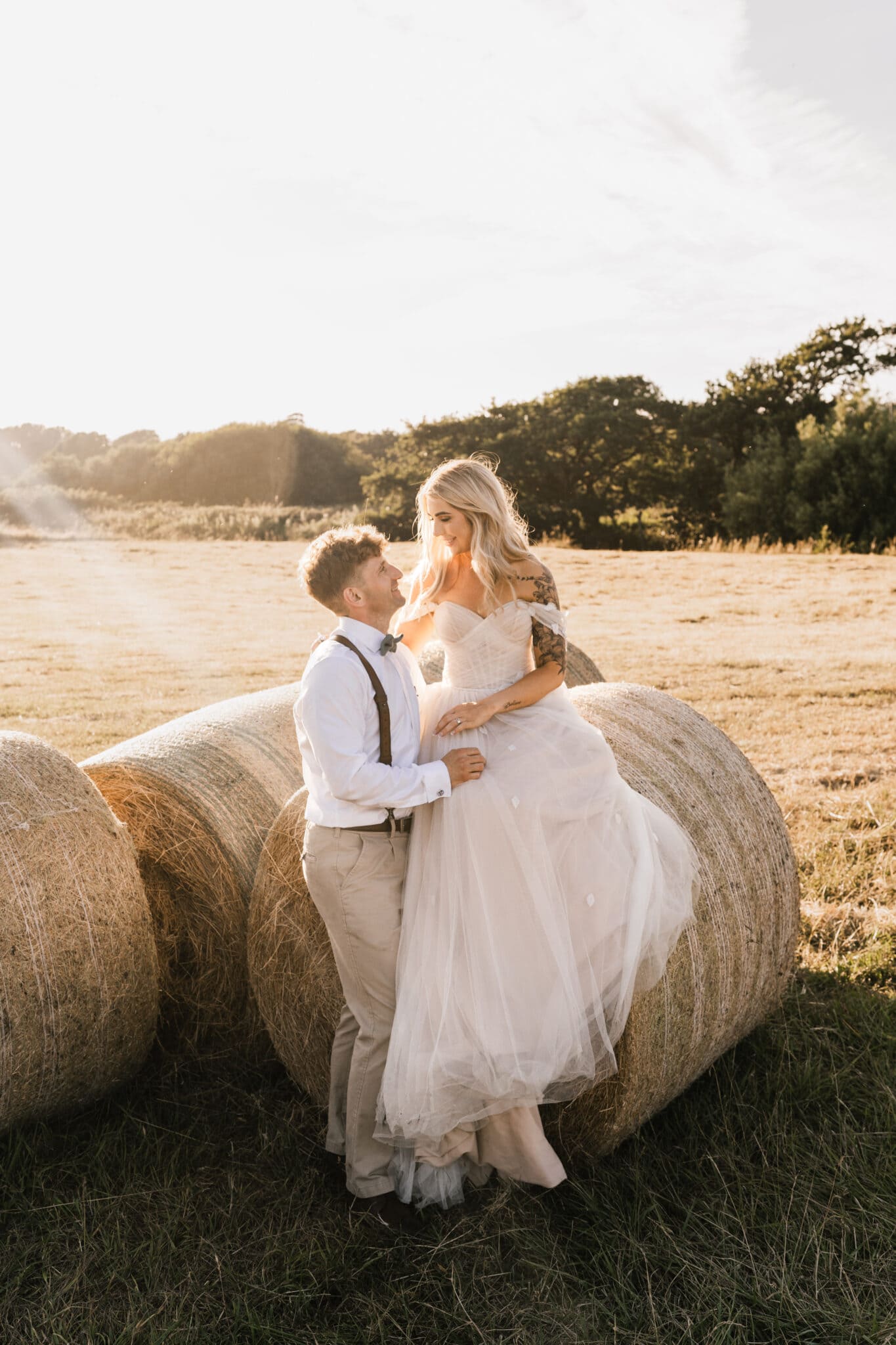 A couple in wedding attire lovingly gazes at each other, standing between large hay bales in a sunlit field. the setting sun casts a warm glow over the scene.