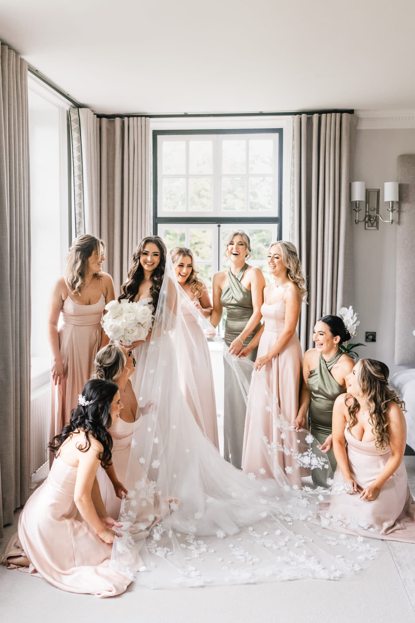 A bride in a white gown with a veil, surrounded by bridesmaids in soft pink dresses, sharing a joyful moment in a stylish room with elegant curtains and soft lighting.