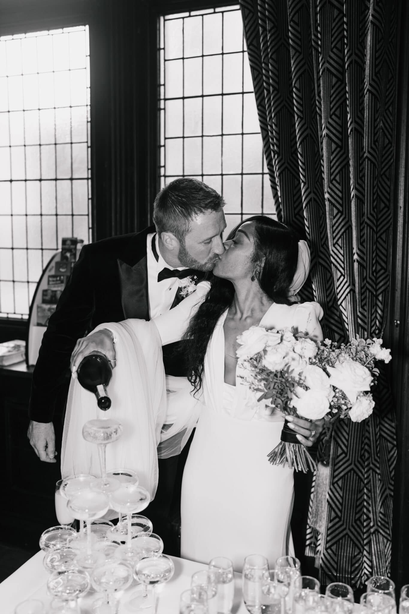 A bride and groom kissing passionately while holding a bridal bouquet, with a background of large windows and patterned curtains in a dimly lit room.
