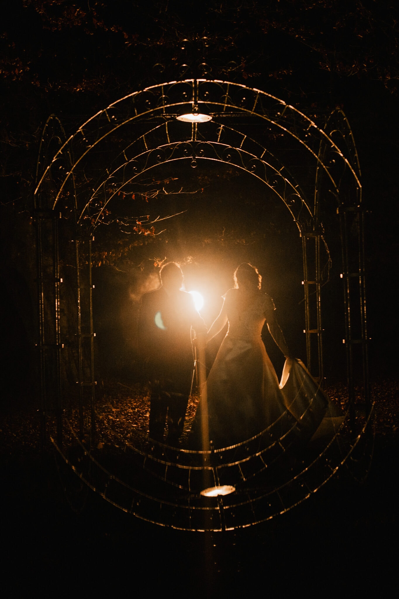 A couple walks through an ornate metal archway at night, illuminated from behind by a bright light that casts a warm glow around them.