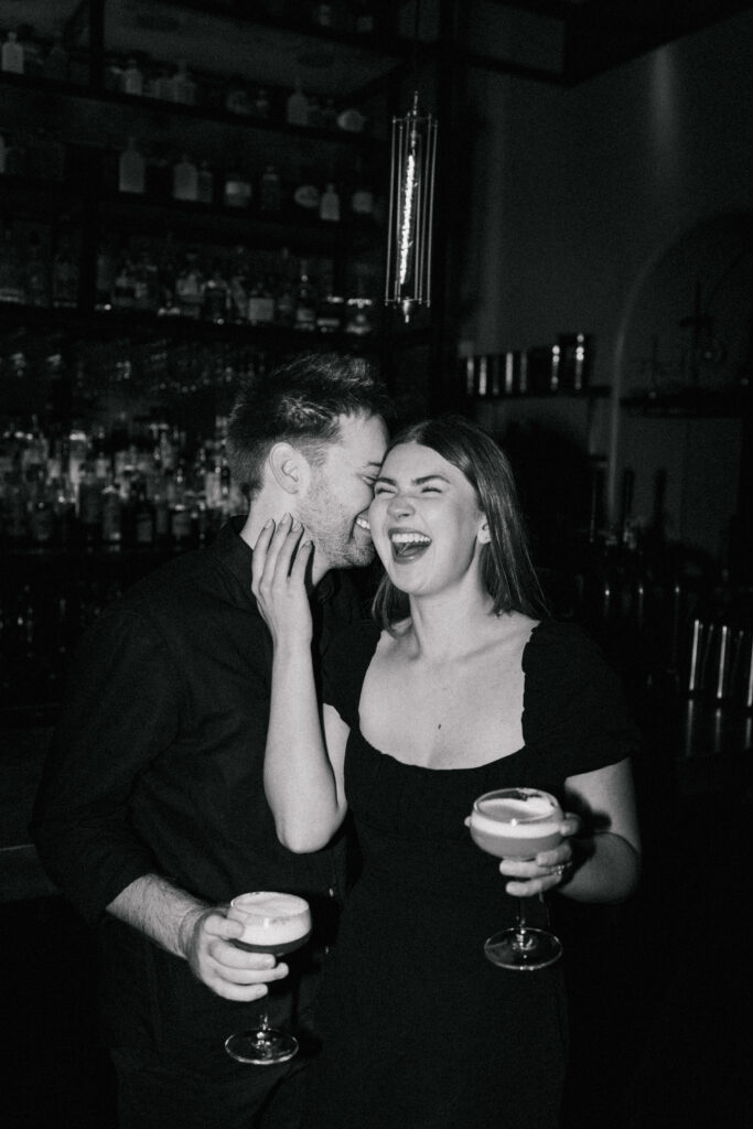 A black and white photo of a smiling couple in a dimly lit bar, each holding a cocktail. The woman laughs joyfully as the man leans in close, their happy expressions conveying a sense of fun and affection.
