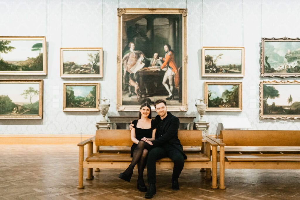 A couple dressed in black sit closely together on a wooden bench in an art gallery, surrounded by framed paintings on the wall behind them, including a large portrait at the centre.