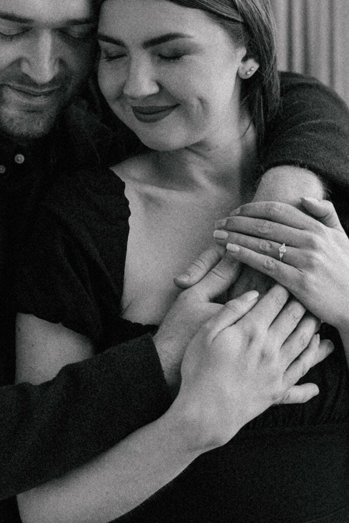 A black and white photo of a smiling woman with closed eyes being embraced from behind. She holds hands with her partner, showing off an engagement ring on her finger.
