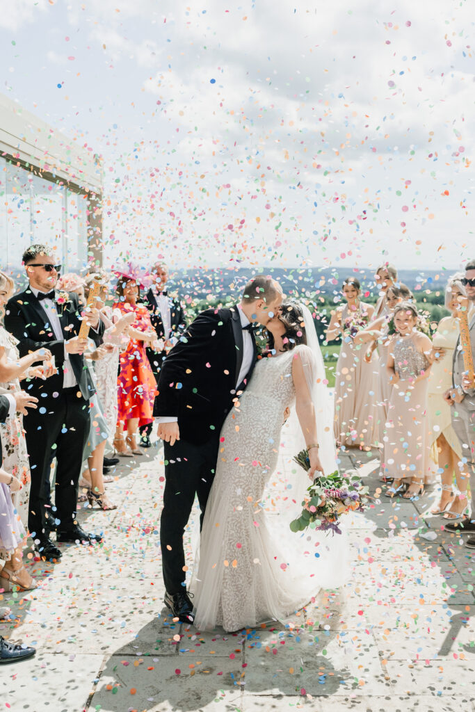 A newlywed couple kiss outdoors surrounded by guests, as colourful confetti rains down. The bride holds a bouquet and wears a white dress and veil; the groom is dressed in a black suit. The crowd smiles and celebrates.