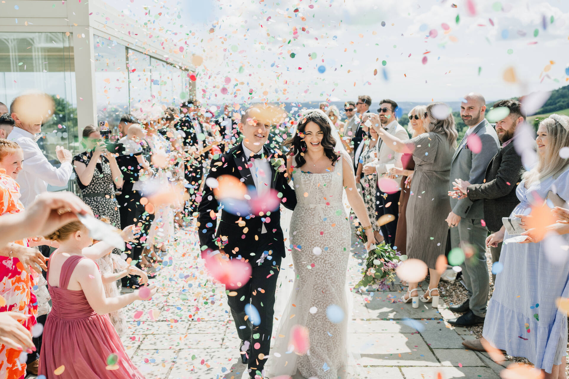 A newlywed couple walks joyfully through a crowd of guests tossing colourful confetti in the air outdoors; the bride wears a white dress and the groom a black tuxedo, both smiling amid the celebration.
