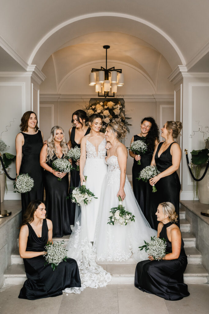 Two brides in white wedding dresses stand close together, surrounded by eight bridesmaids in black dresses holding bouquets. They are all smiling and posing on a staircase under an elegant chandelier.