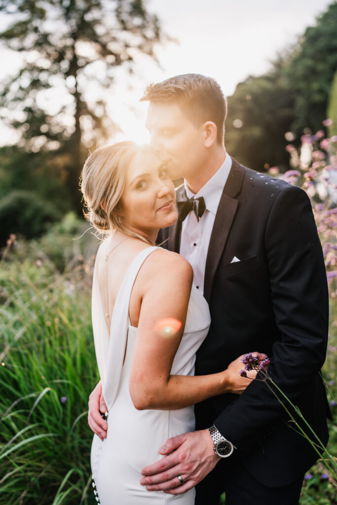 A bride in a white dress and groom in a black tuxedo embrace outdoors at sunset, surrounded by greenery and wildflowers. Captured by a South Wales Wedding Photographer, the groom kisses the bride’s forehead whilst she looks at the camera.