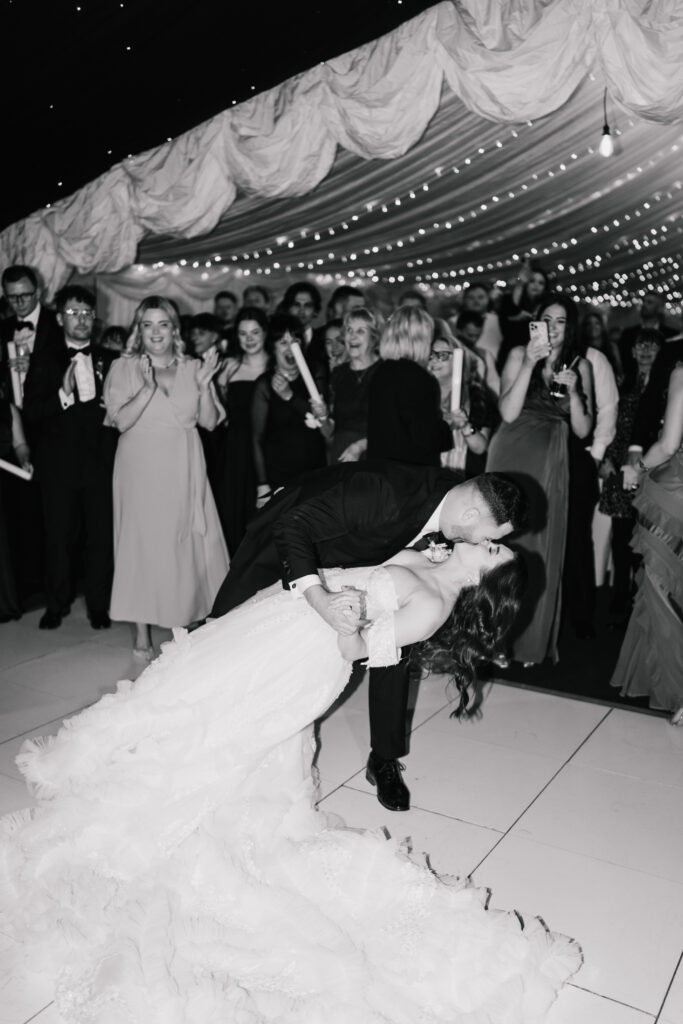 A groom dips and kisses his bride on the dance floor as a crowd of guests, dressed formally, watches and cheers in a decorated, draped venue with fairy lights. The moment is joyful and celebratory.