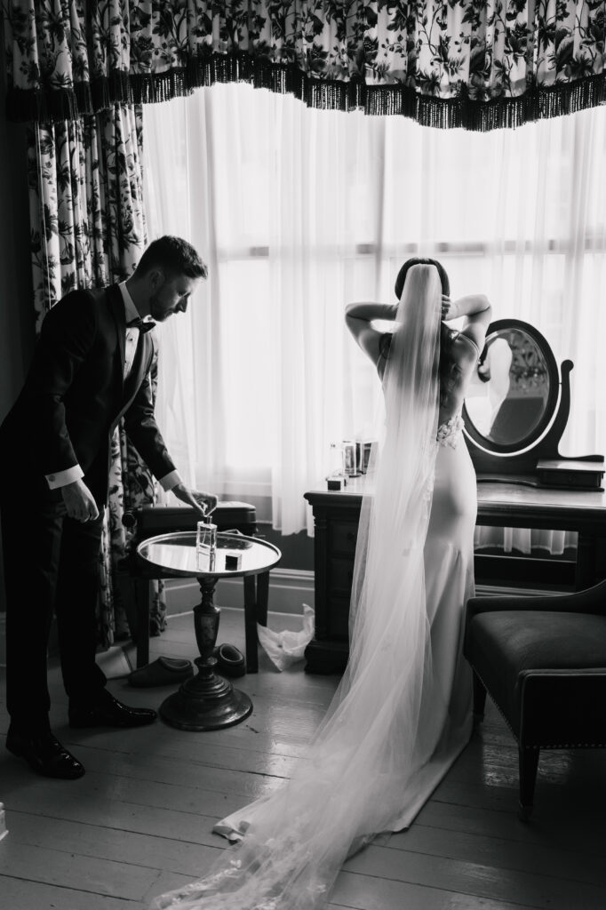 A groom in a suit pours a drink at a small table while a bride in a long veil adjusts her hair at the dressing table, all captured in soft light and floral elegance—an intimate moment showcasing timeless wedding photography style.