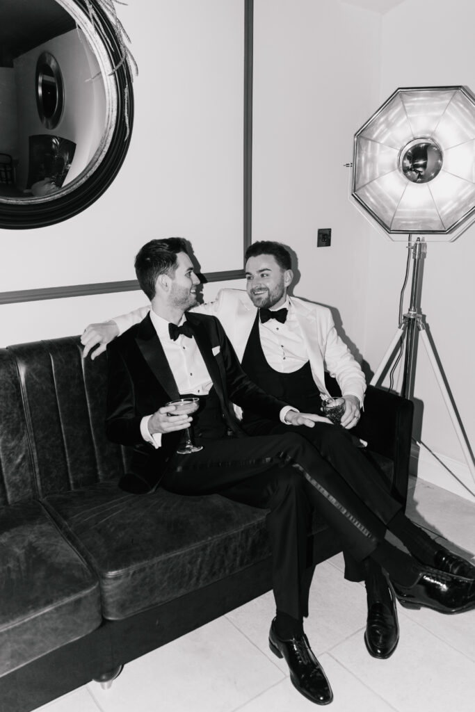 Two men in tuxedos sit on a black sofa, smiling at each other and holding drinks. Styled with a Wedding Photography Style touch, a round mirror and large studio light accent the modern black-and-white room.