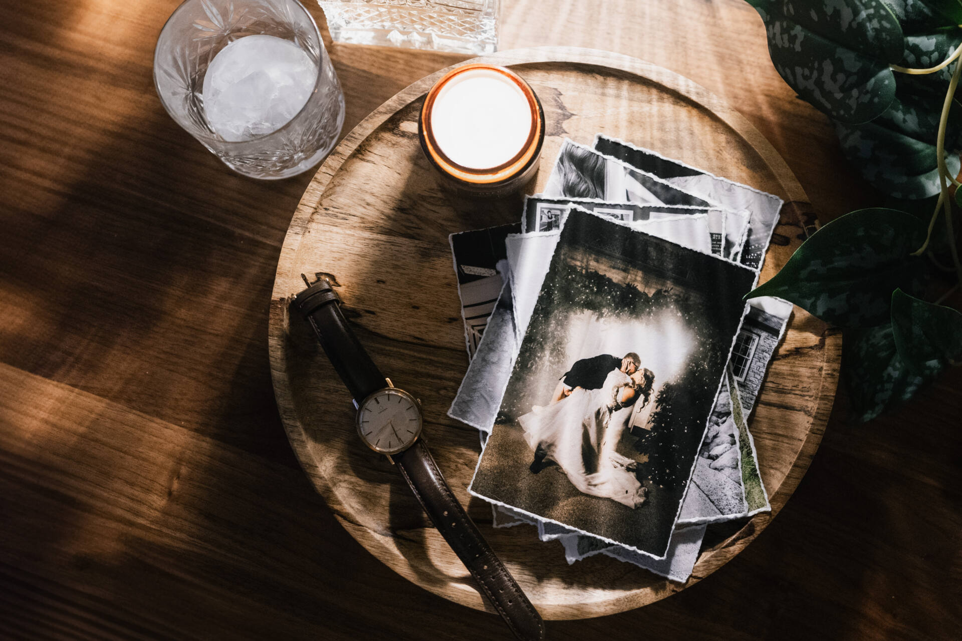 A wooden tray holds a stack of black-and-white and colour photos, a lit candle, a wristwatch with a brown strap, and a glass of iced water, with sunlight streaming across a wooden table and green leaves nearby.