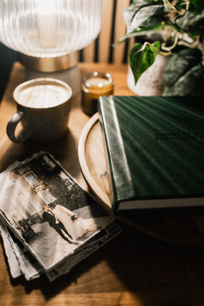 A cosy scene with a cup of coffee, a lit candle, a potted plant, a green photo album, and a stack of black-and-white wedding photos on a wooden table under a lamp.