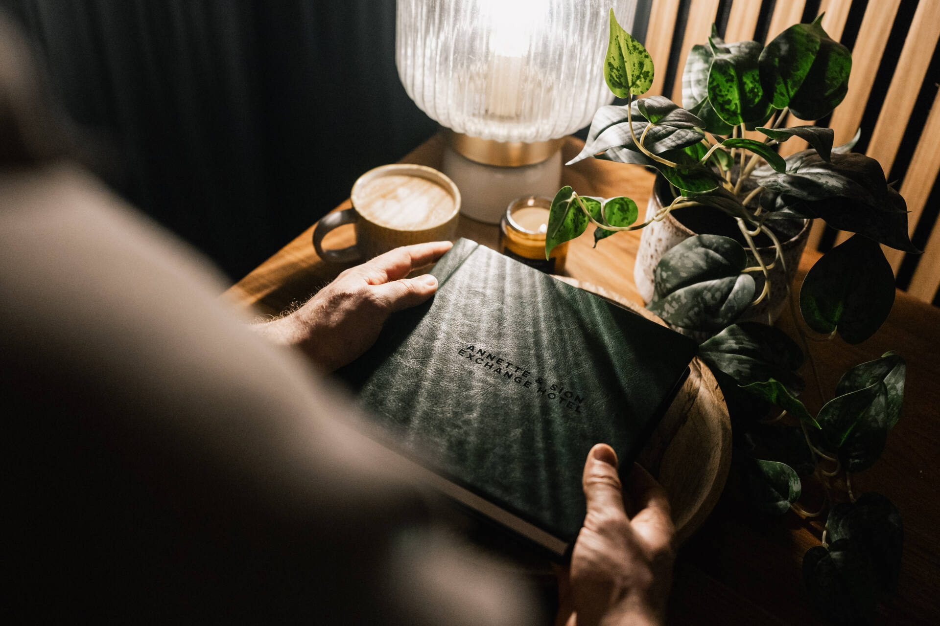 A person holds a closed black book at a wooden table with a lamp, a potted plant, a cup of coffee, and a small candle. Warm light creates a cosy atmosphere.