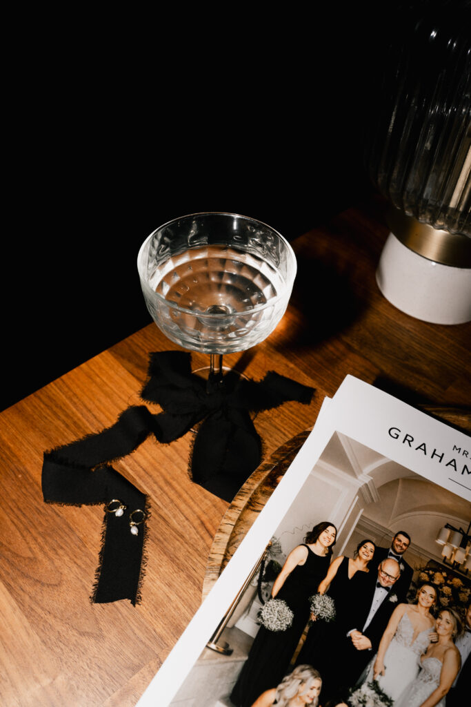 A cocktail glass with a black ribbon sits on a wooden table next to black fabric swatches with earrings and a wedding photo booklet featuring people in formal attire. A lamp is partially visible in the background.
