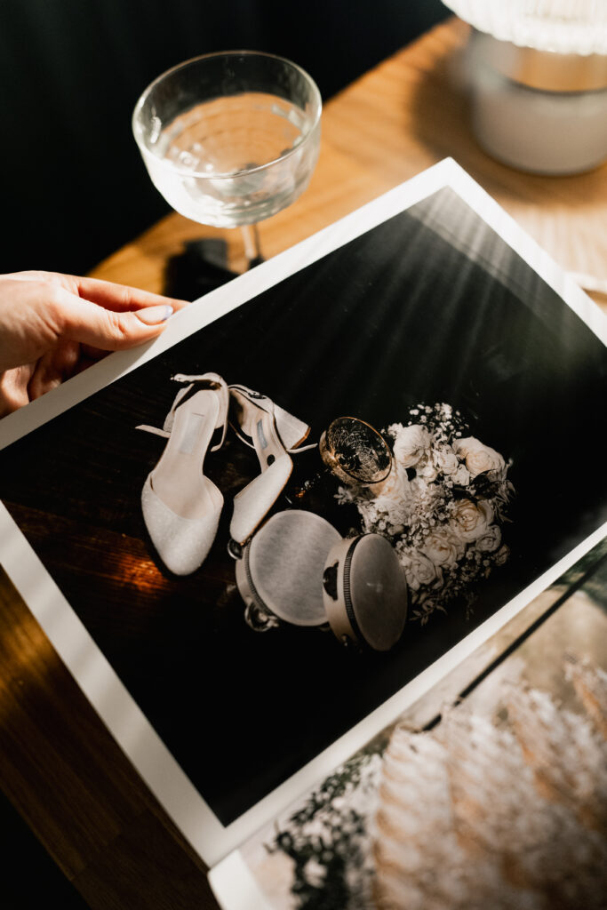 A hand holds open a photo album on a wooden table, displaying a photo of white high-heeled shoes, a gold-rimmed glass, and a bouquet of white flowers; a glass and lamp are also visible on the table.