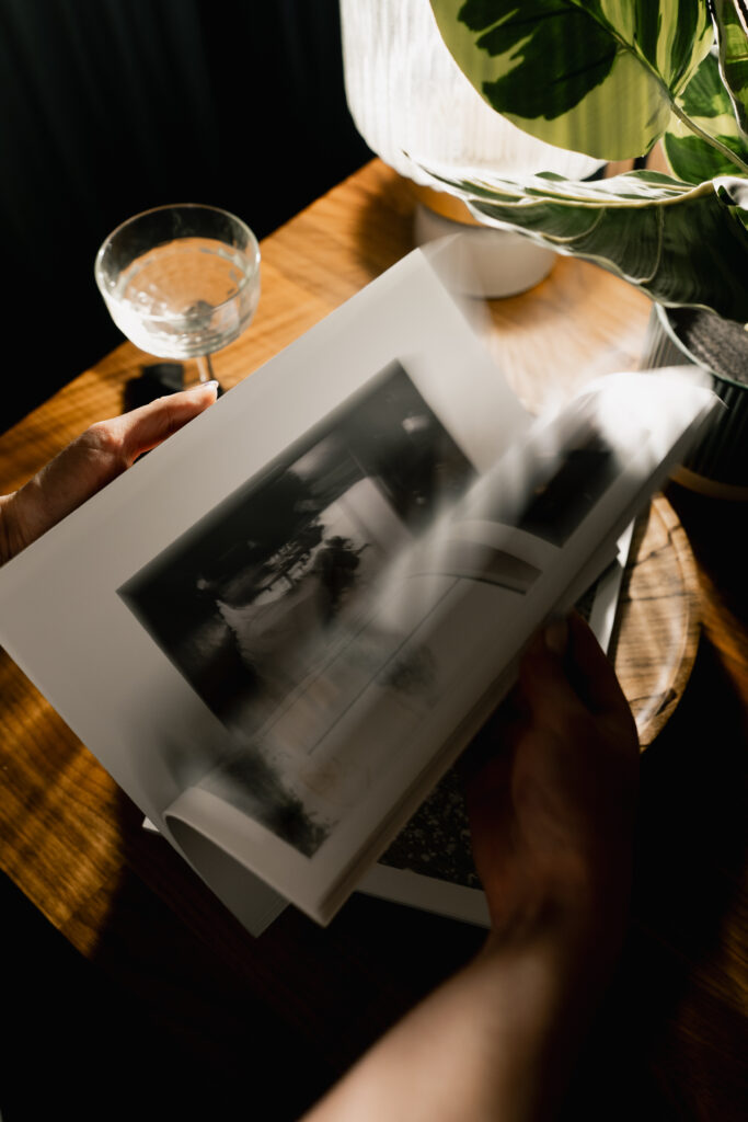A person flicks through a photo book on a wooden table next to a potted plant, a lamp, and a glass with a drink, with sunlight casting shadows across the scene.