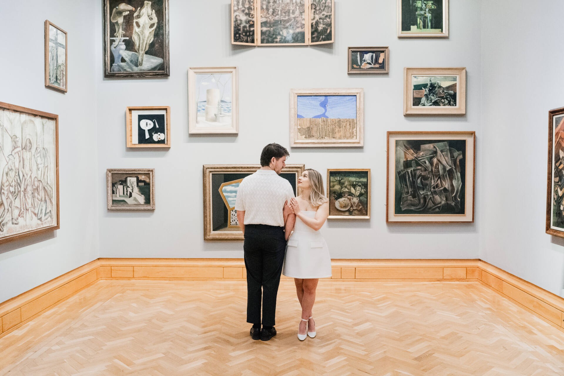 A couple stands in the centre of an art gallery, holding hands and looking at each other, surrounded by framed paintings—capturing a timeless moment perfect for a wedding photo album.