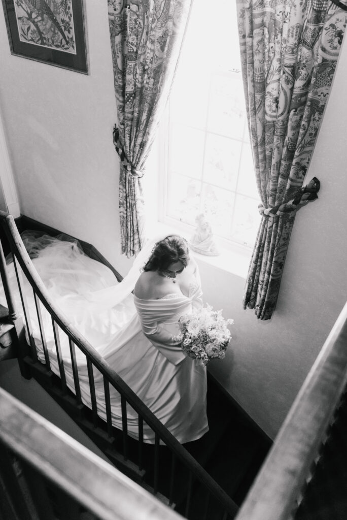 A bride in a flowing gown and veil holds a bouquet whilst sitting on a staircase by a bright window with patterned curtains, bathed in natural light. Captured by a South Wales Wedding Photographer, the timeless image is in black and white.