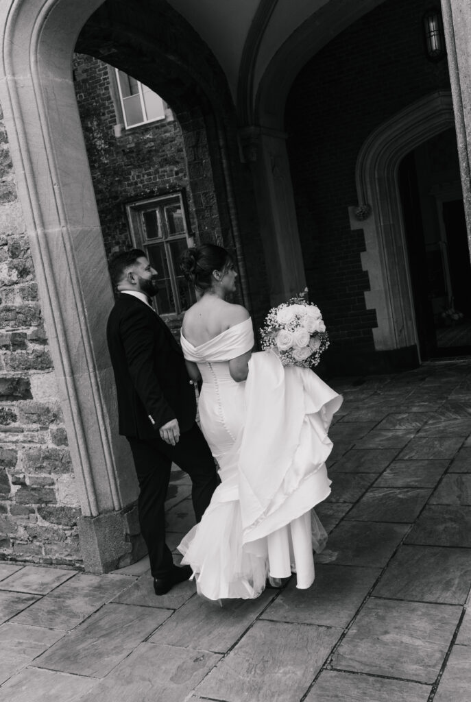 A bride and groom walk under a stone archway in this black and white scene, perfect for a timeless wedding photography album. The bride’s white gown and the groom’s suit stand out against the elegant architectural backdrop.