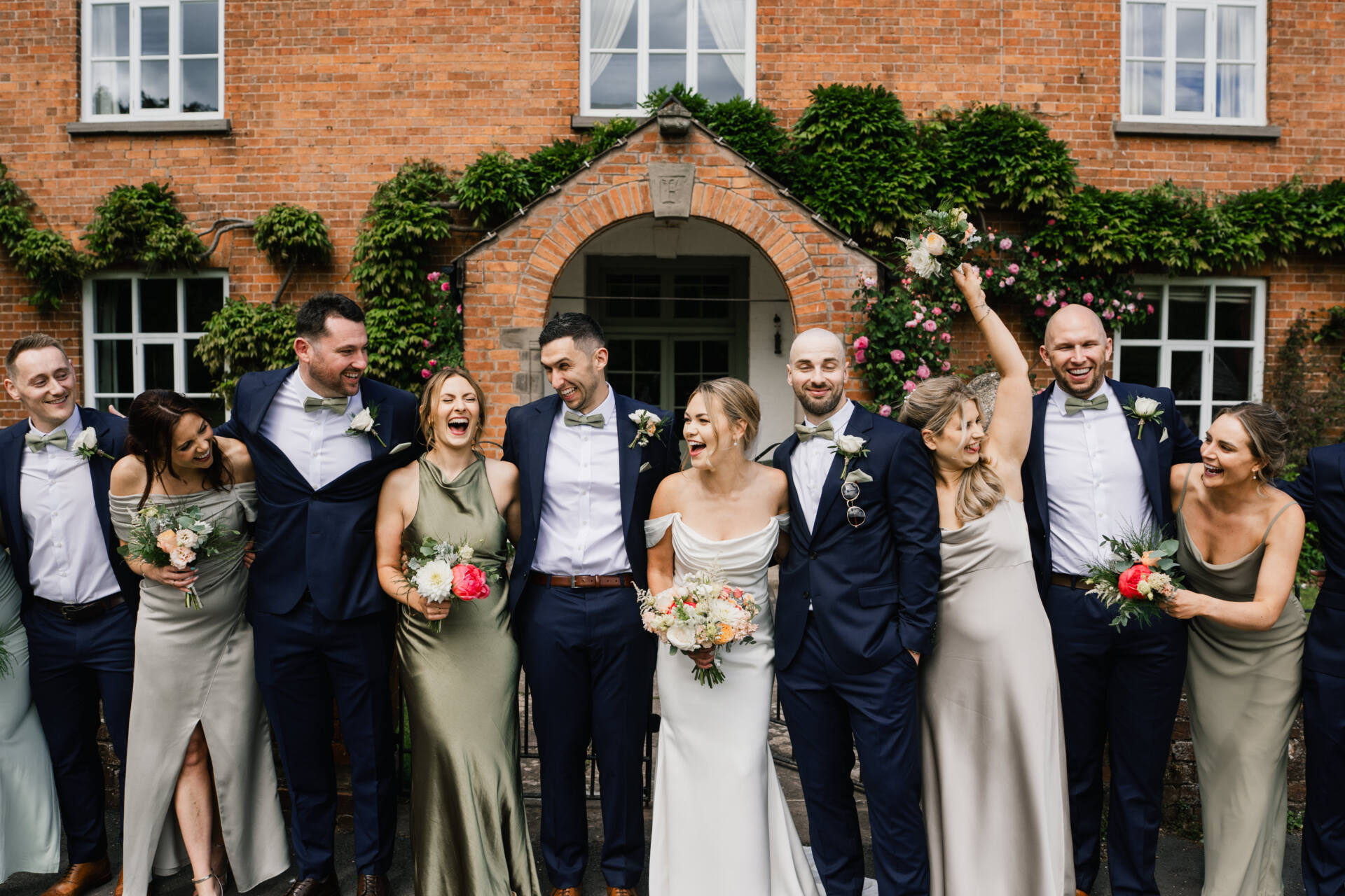 A wedding party stands in front of a brick building, smiling and laughing. Perfect for a wedding photo album, the bride and groom are in the centre, surrounded by bridesmaids in sage green dresses and groomsmen in navy suits.