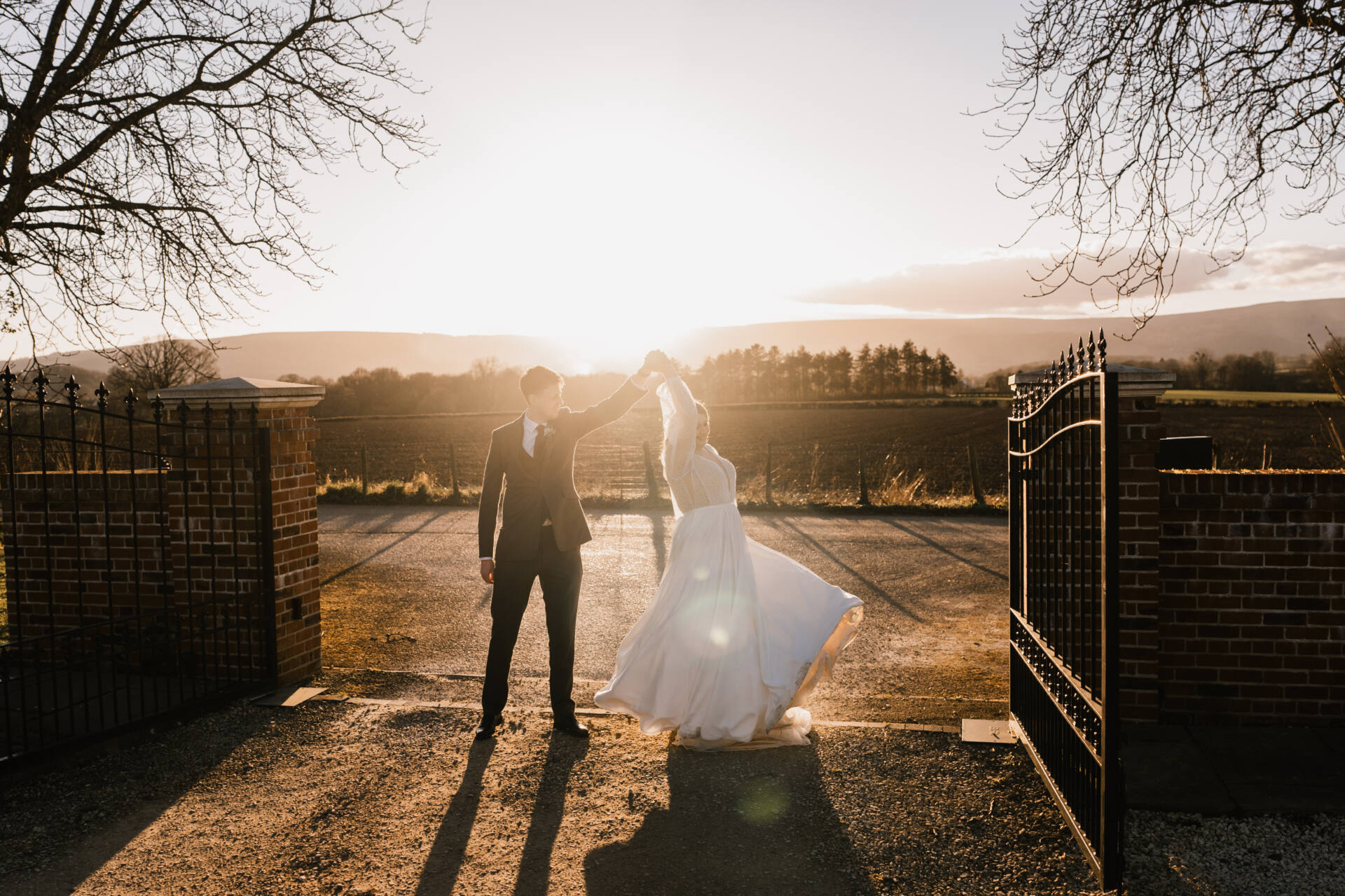 A bride and groom dance at sunset near an open iron gate, with the sun shining behind them and trees framing the scene. The bride’s dress flows as she twirls, and the landscape is bathed in warm light.