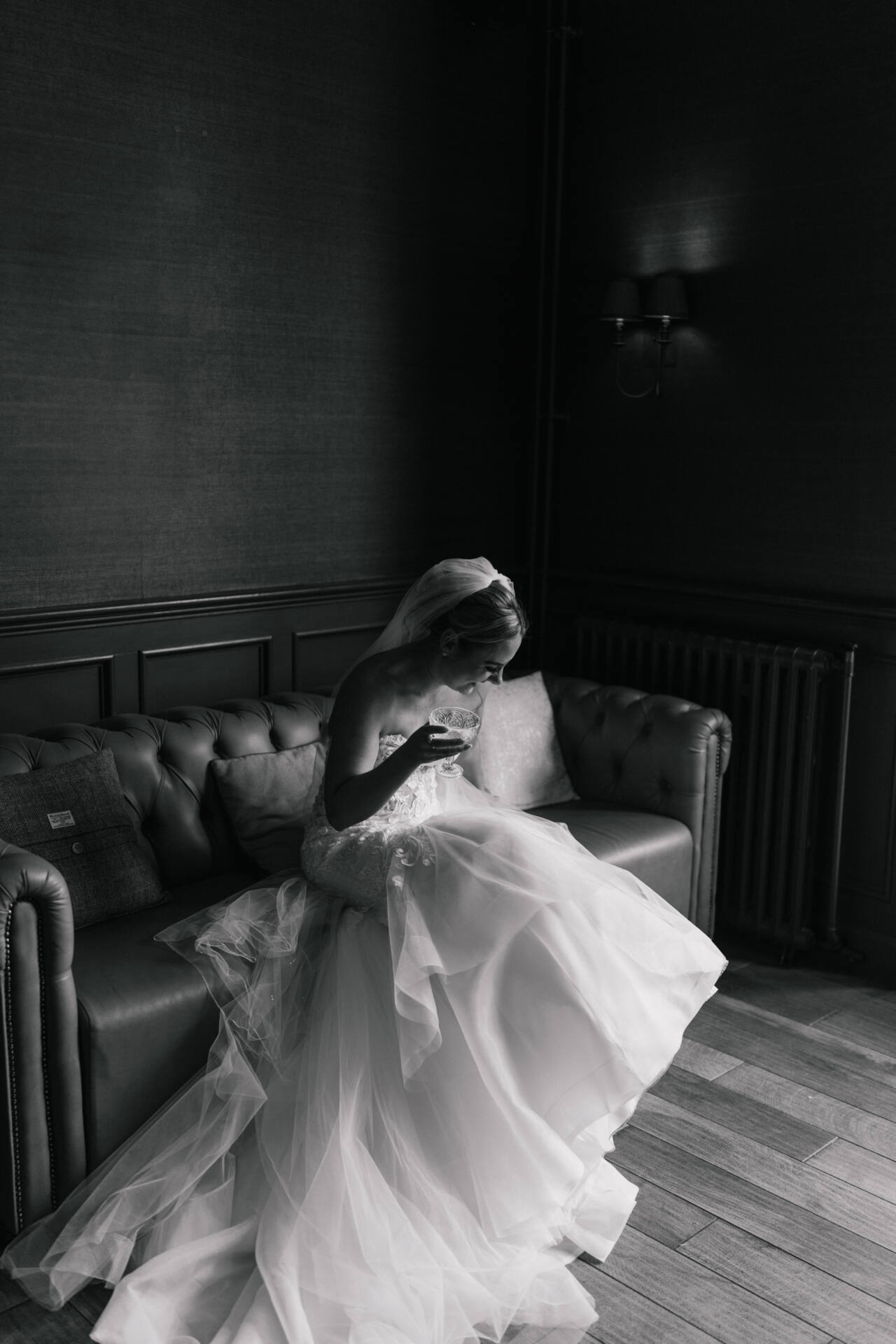 A bride in a wedding dress sits alone on a sofa, looking down at her bouquet in a dimly lit room. Captured by a Bryngarw House Wedding Photographer, the black and white photo creates a calm and contemplative mood.